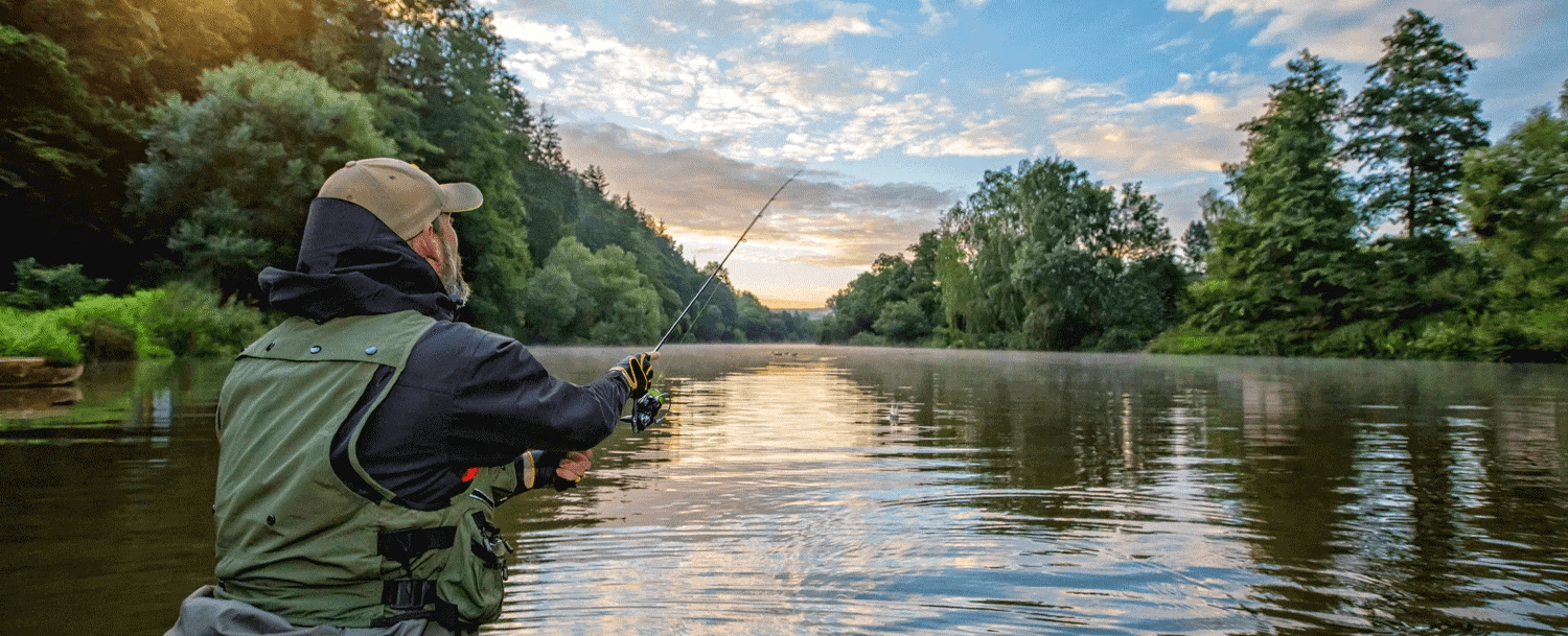 A fisherman stands in a boat casting his line on a tranquil river at dawn.