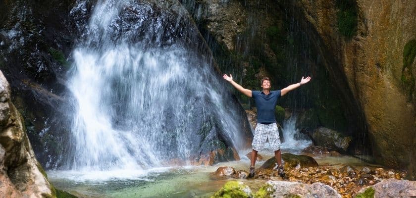 A person stands with arms outstretched in front of a cascading waterfall.