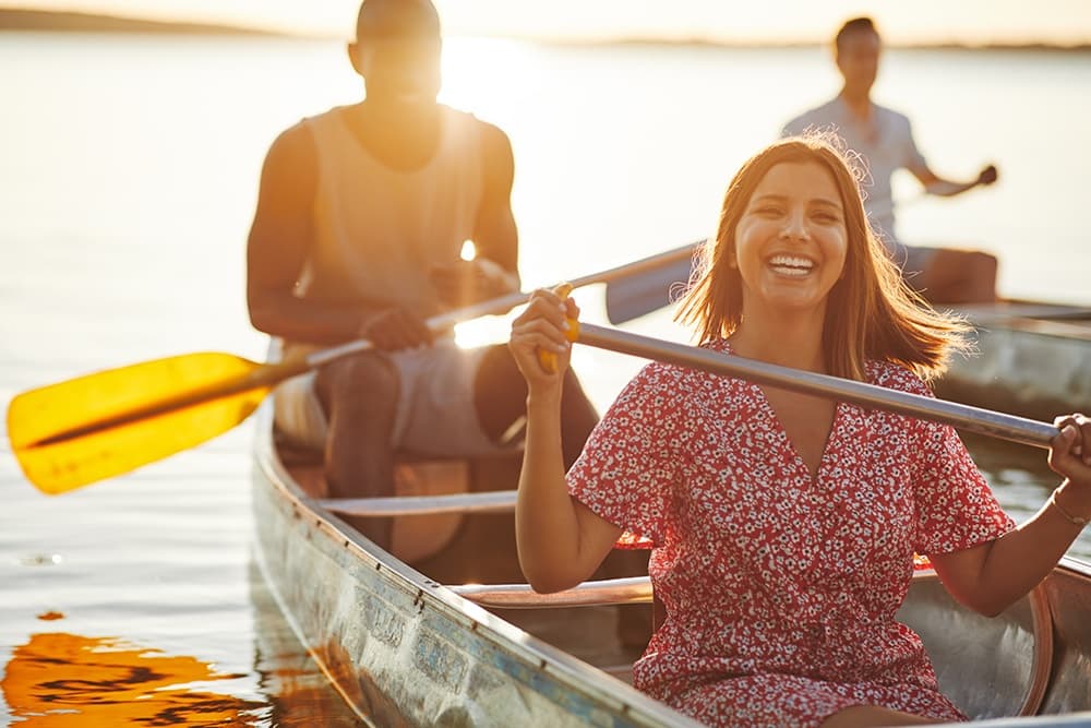 A joyful woman in a floral dress rows a canoe on a lake, with two companions in the background.