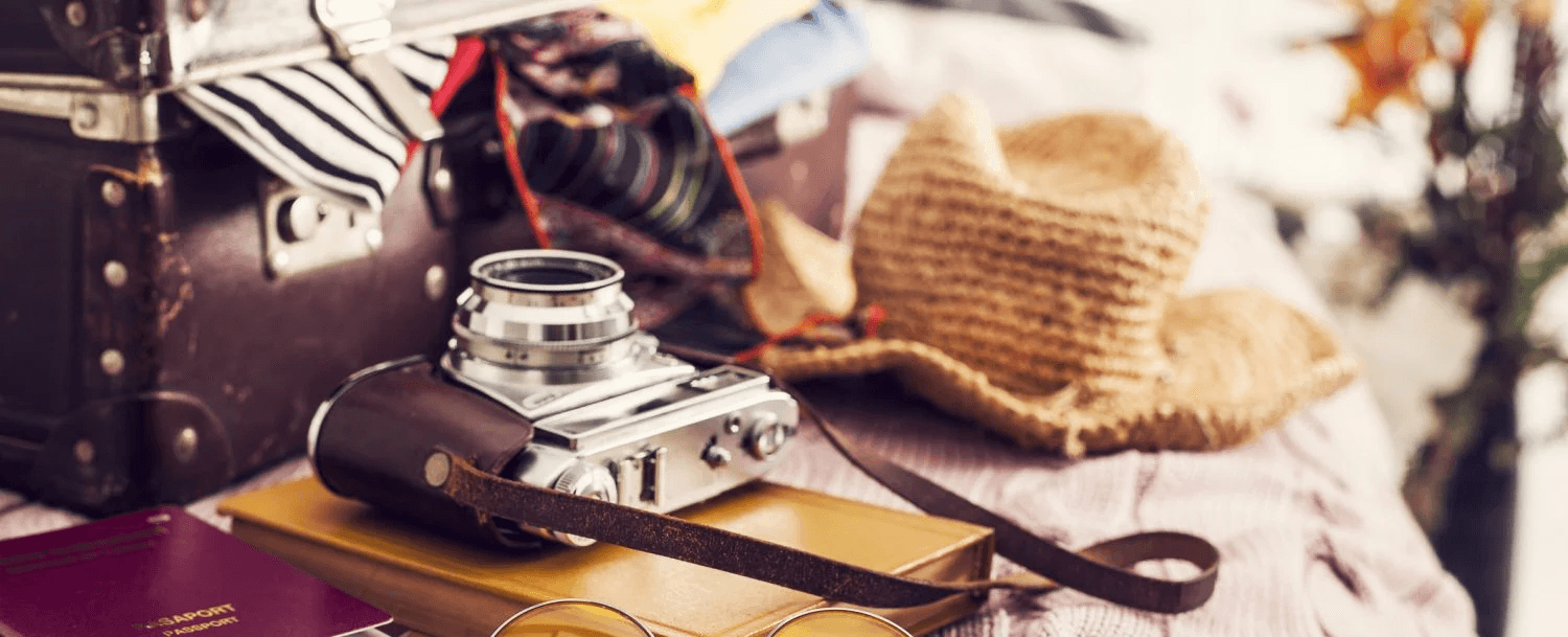 A vintage camera, passport, and glasses are placed on a yellow notebook beside a straw hat and an open suitcase filled with clothing.