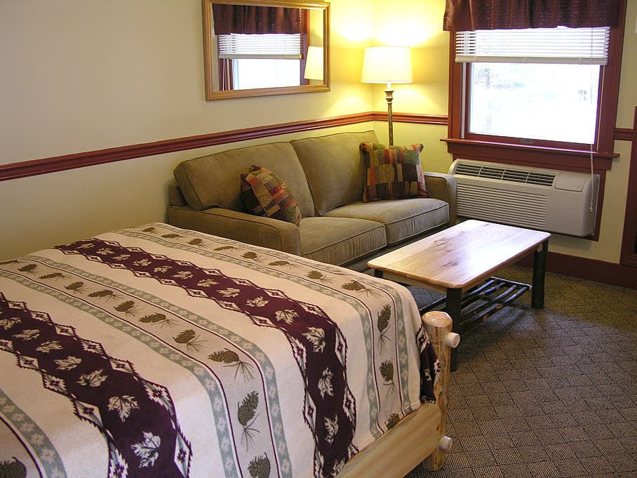 A light-filled room with a queen bed and a tan sofa separated by a wooden coffee table against a wall with red trim.