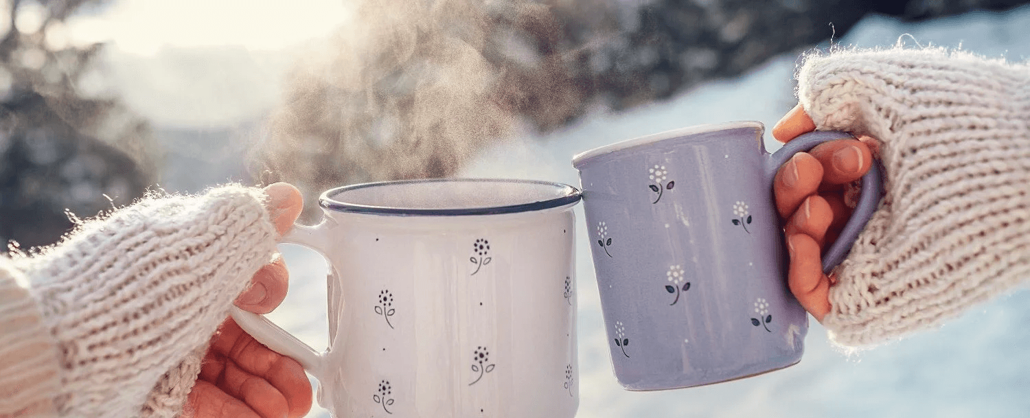 Two hands in cozy mittens hold steaming mugs in a snowy landscape.