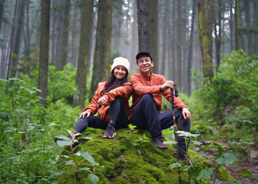 Two people in matching orange jackets sit on a mossy rock in a lush forest. Two people in matching orange jackets sit on a mossy rock in a lush forest.