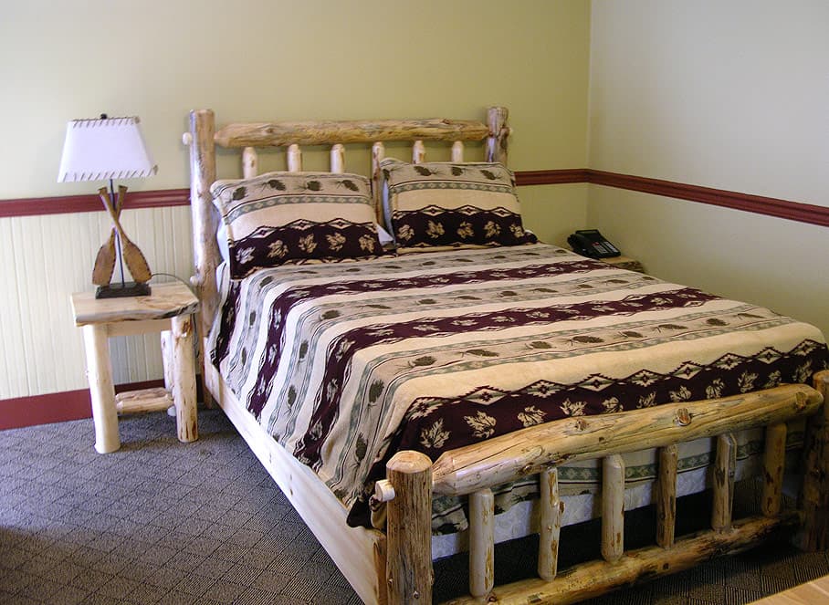 A rustic bedroom featuring a log-style queen bed with a striped, patterned quilt, a matching log nightstand, and a paddle lamp.