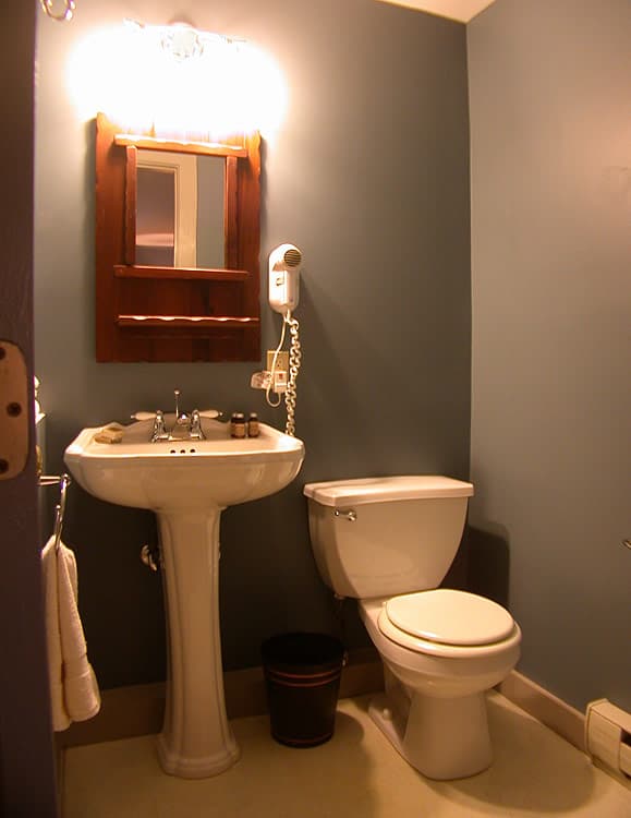 A small bathroom with a white pedestal sink, a toilet, a wooden-framed mirror, and a wall-mounted hairdryer against a blue wall.