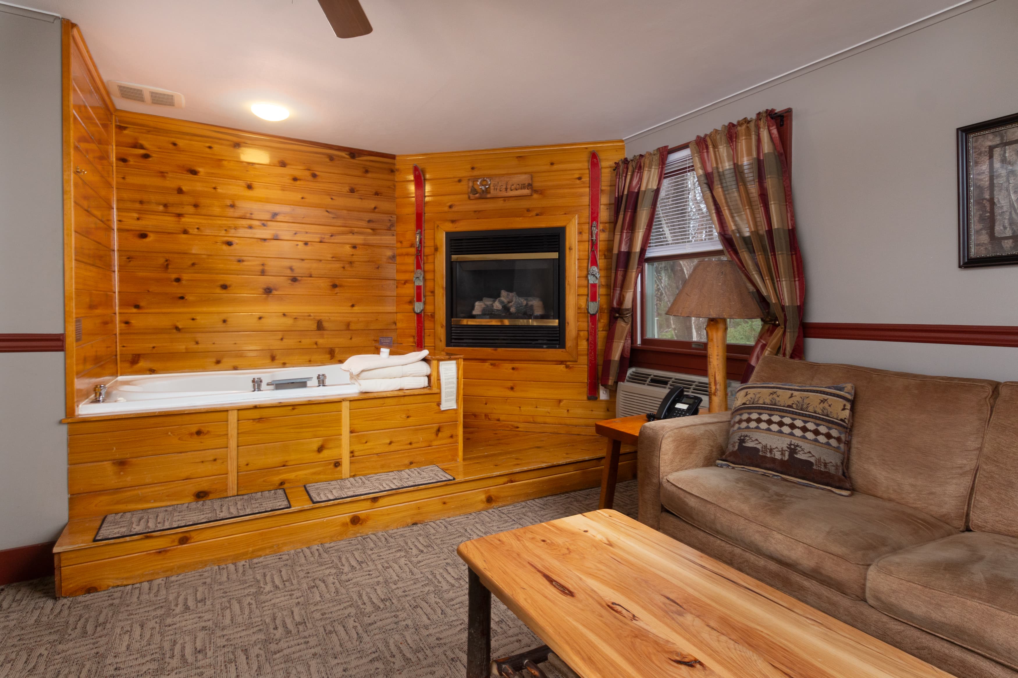 A rustic suite's sitting area featuring a tan sofa, a wooden coffee table, and a corner built-in area with a jetted tub and a fireplace.