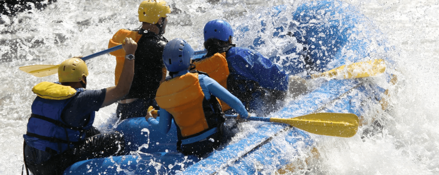 A group of four people paddles a blue raft through choppy waters, splashing water everywhere.