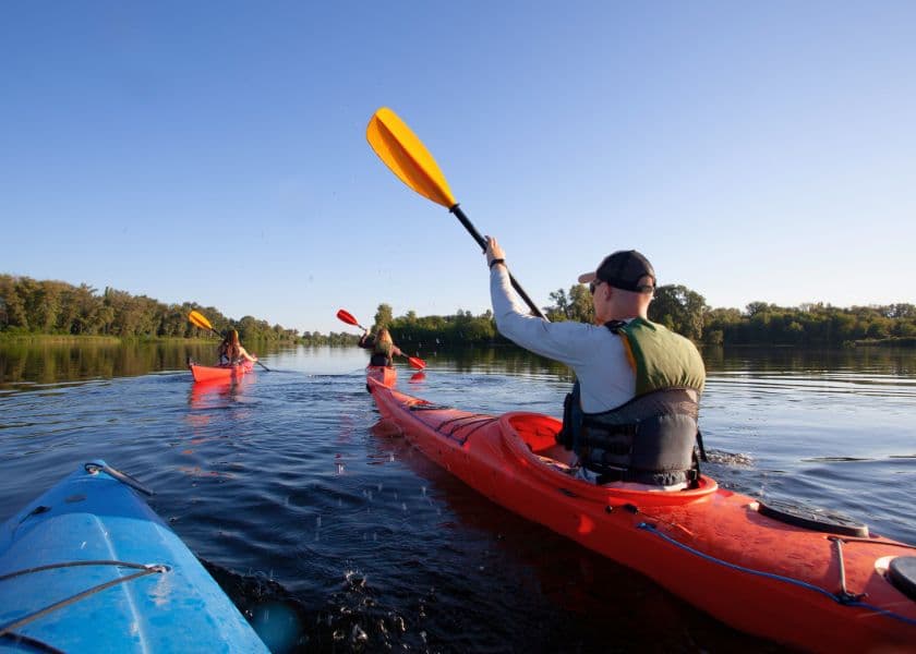 Three kayakers paddle on a calm river under a clear blue sky. Three kayakers paddle on a calm river under a clear blue sky.