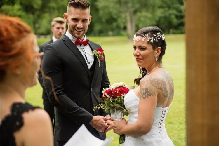 A smiling bride and groom hold hands during their wedding ceremony in a lush outdoor setting.
