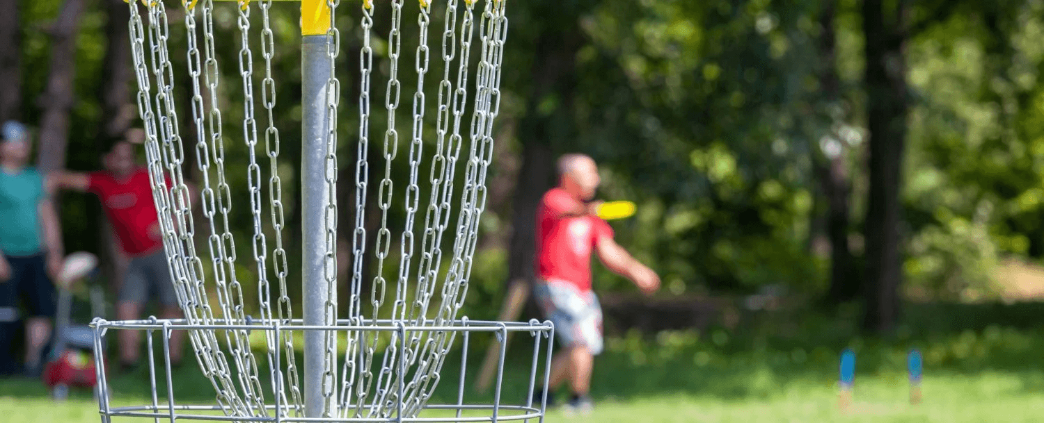 A disc golf basket is in focus with players throwing discs in the background.