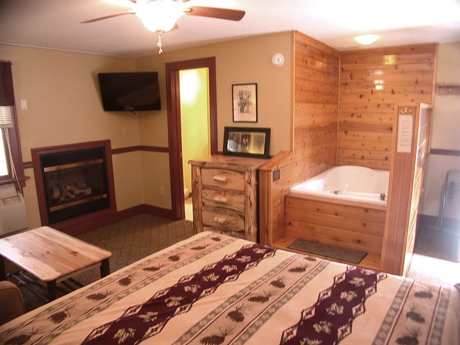 A rustic suite with a queen bed, a built-in fireplace and TV, and a jetted tub in a wood-paneled corner.
