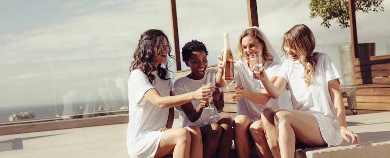 Four women in white t-shirts celebrate with drinks on a sunny patio. Four women in white t-shirts celebrate with drinks on a sunny patio.