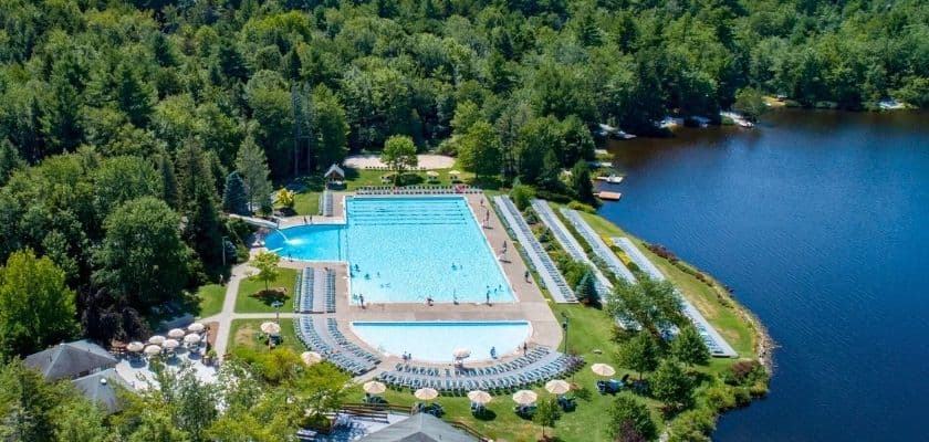 Aerial view of a large outdoor swimming pool beside a lake, surrounded by lush trees.