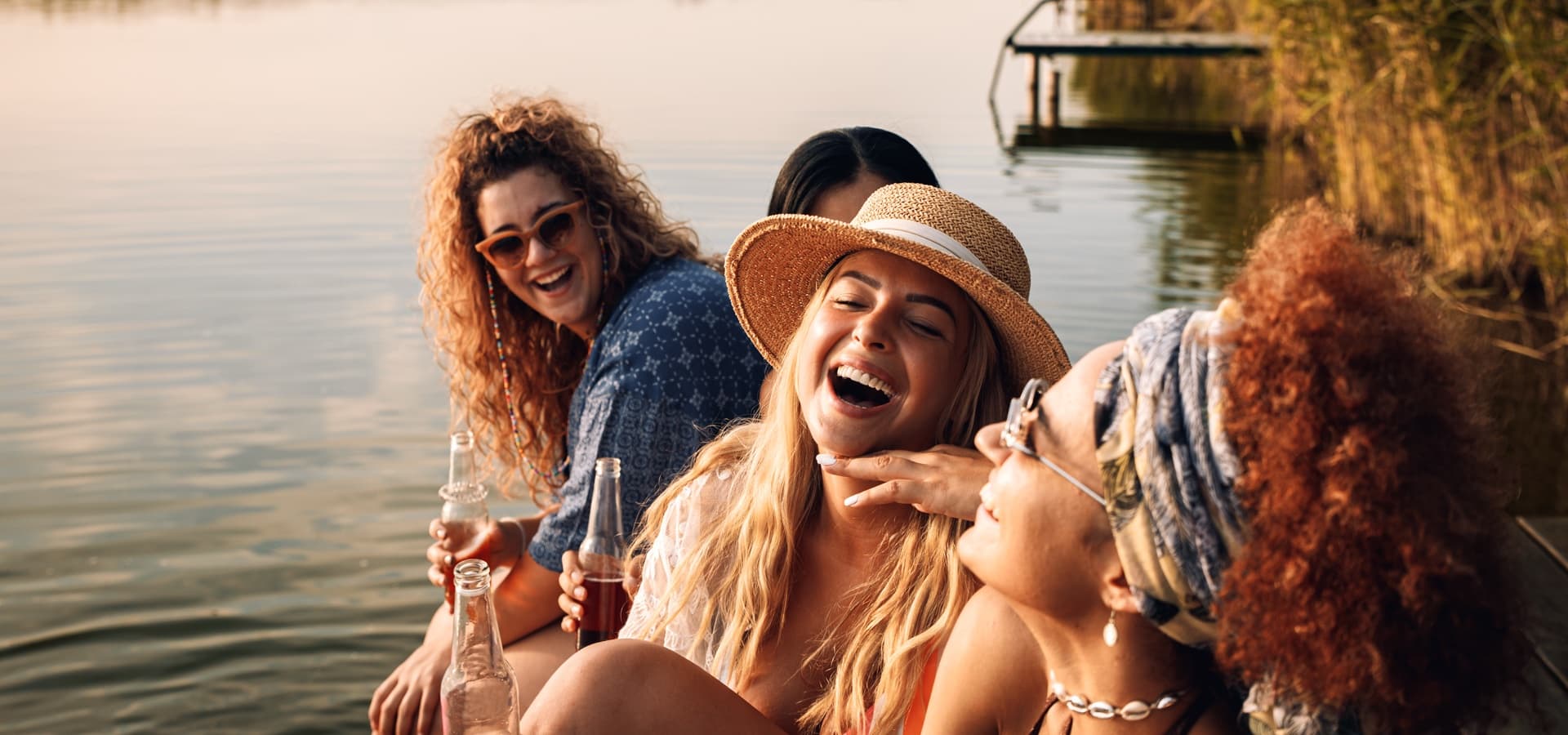 A group of four women joyfully laughing while sitting by a calm lake during sunset.