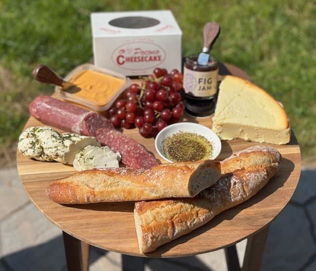 A wooden table displays an assortment of cheeses, cured meats, baguettes, grapes, fig jam, and a cheesecake.