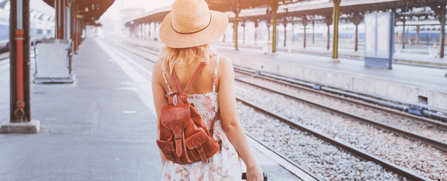 A woman in a floral dress and hat stands at a train station, facing the railway tracks.