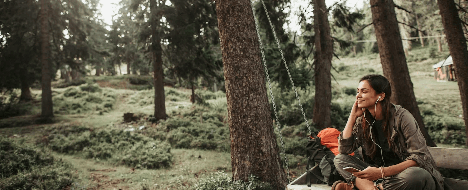 A woman sits in a forest, wearing headphones and looking thoughtfully at her phone.