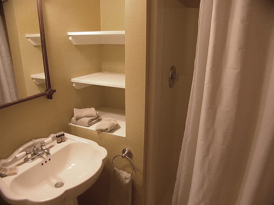 A bathroom with a white pedestal sink, a shower with a white curtain, and recessed built-in shelving on a pale yellow wall.