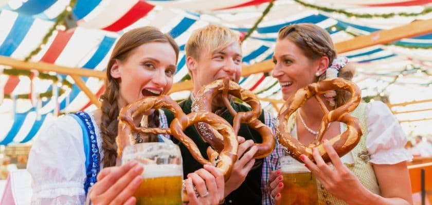 Three friends in traditional attire joyfully hold pretzels and beers under a colorful tent.