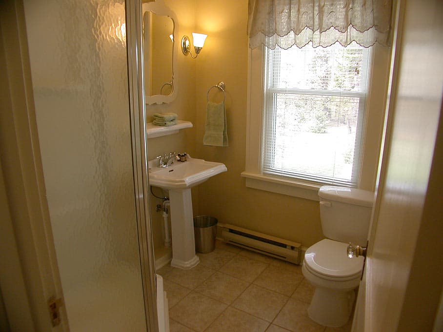 A small, bright bathroom with a pedestal sink, a toilet under a window with a lace valance, and a glass-door shower.