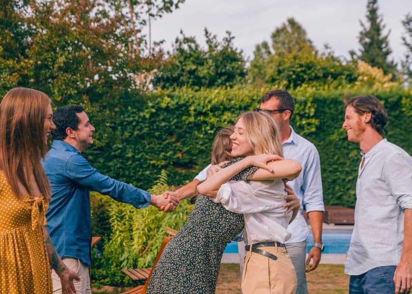 a group of friends meeting in a backyard near a pool warmly hugging and shaking hands surrounded by trees and outdoor furniture
