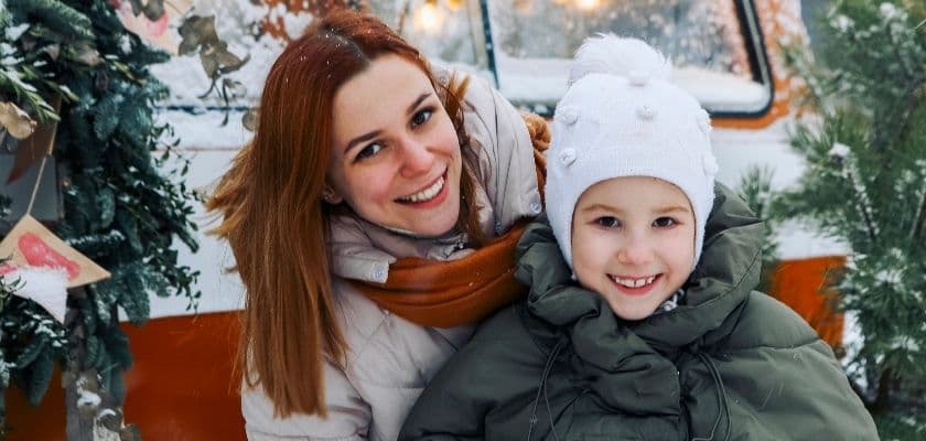 a mother and child dressed in warm winter coats and hats smile in a snowy holiday setting