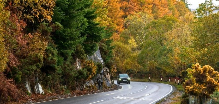 A car driving on a winding road through a forest of autumn foliage.